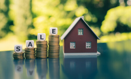 Concept for a house on sale. A hand holds a model house above a meadow. Dice form the word "sale".